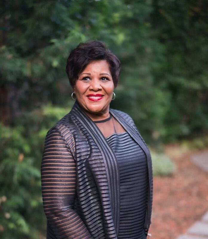 Smiling woman wearing a sheer black jacket and striped top, standing outdoors with greenery in the background.