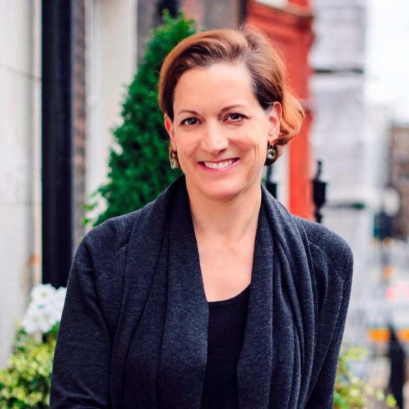 A person with short auburn hair is smiling, wearing a dark cardigan, standing outdoors near greenery and buildings in the background.