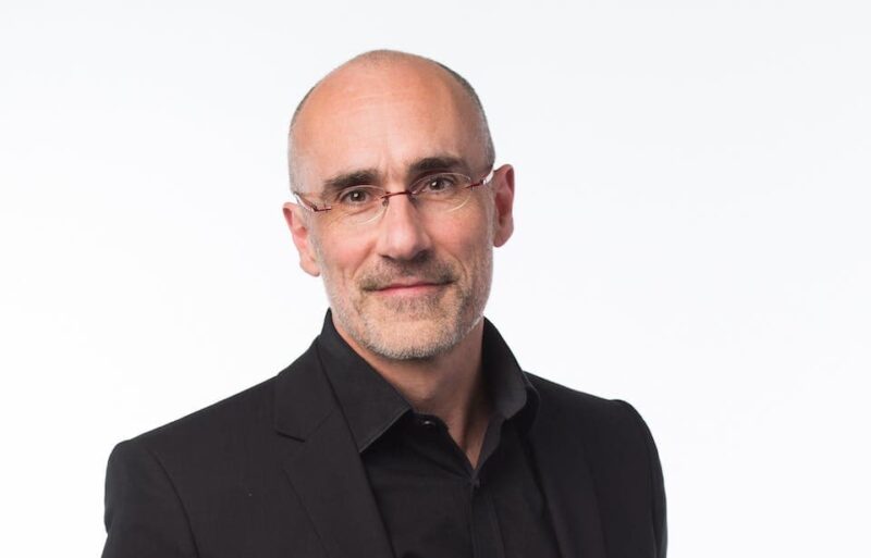 Man with glasses, bald on top with short hair on sides, wearing a black blazer and shirt, looking at the camera against a plain white background.