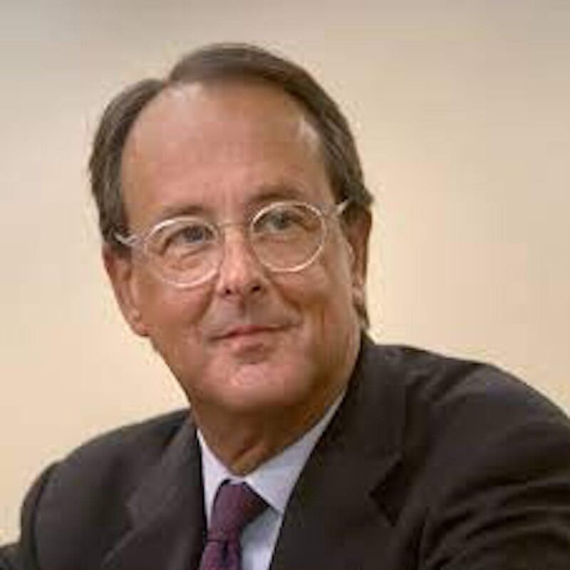 Man with glasses and a suit, looking to the side with a slight smile. Neutral beige background.