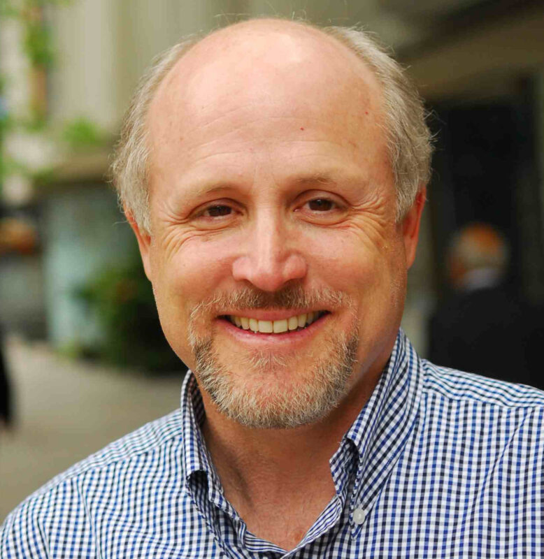 A smiling man with a goatee wearing a checkered shirt stands outdoors.