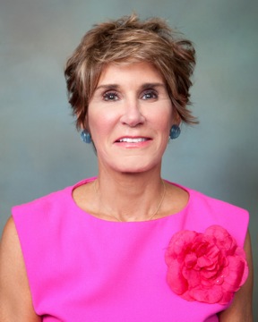 Person in a bright pink top with a flower detail, short brown hair, and blue earrings, smiling against a blurred green background.