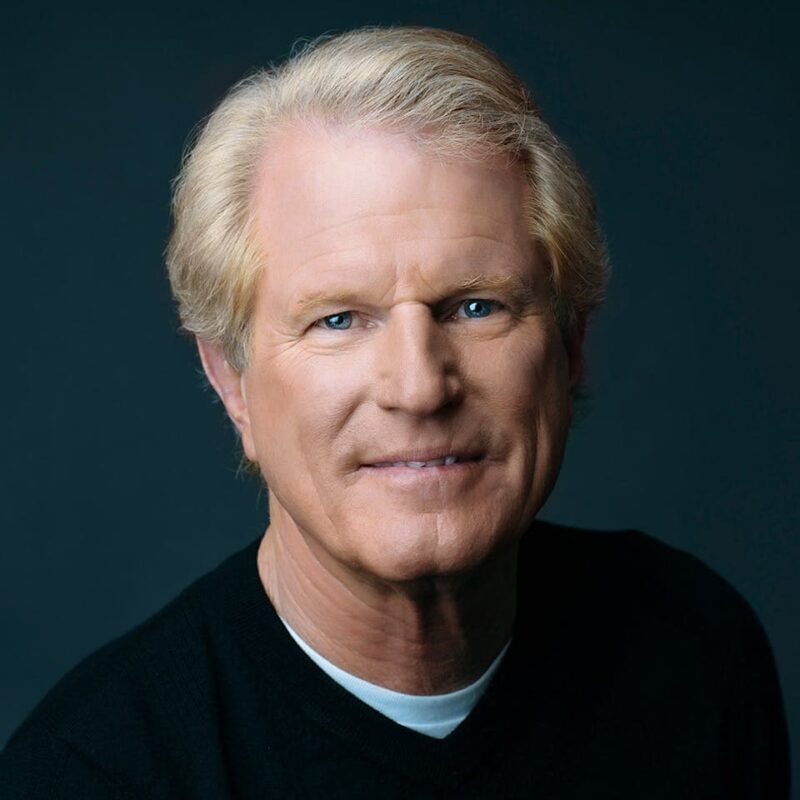 Person with short blonde hair wearing a black shirt, smiling slightly against a dark background.