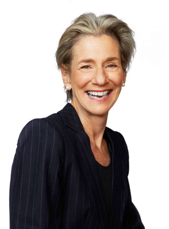 A smiling woman with short, light hair wears a black pinstripe blazer against a white background.