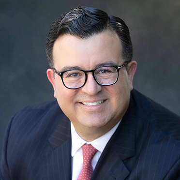 Smiling man in glasses wearing a pinstripe suit and red tie, with dark hair, against a dark background.